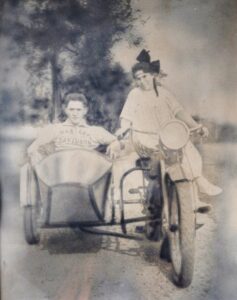 Black and white photo of a woman sitting on an old fashioned motorbike with a man riding sidecar, the image is from 1970 and depicts Ovila and Leone Cadorette, who owned the farmland that later became NCC's campus. 